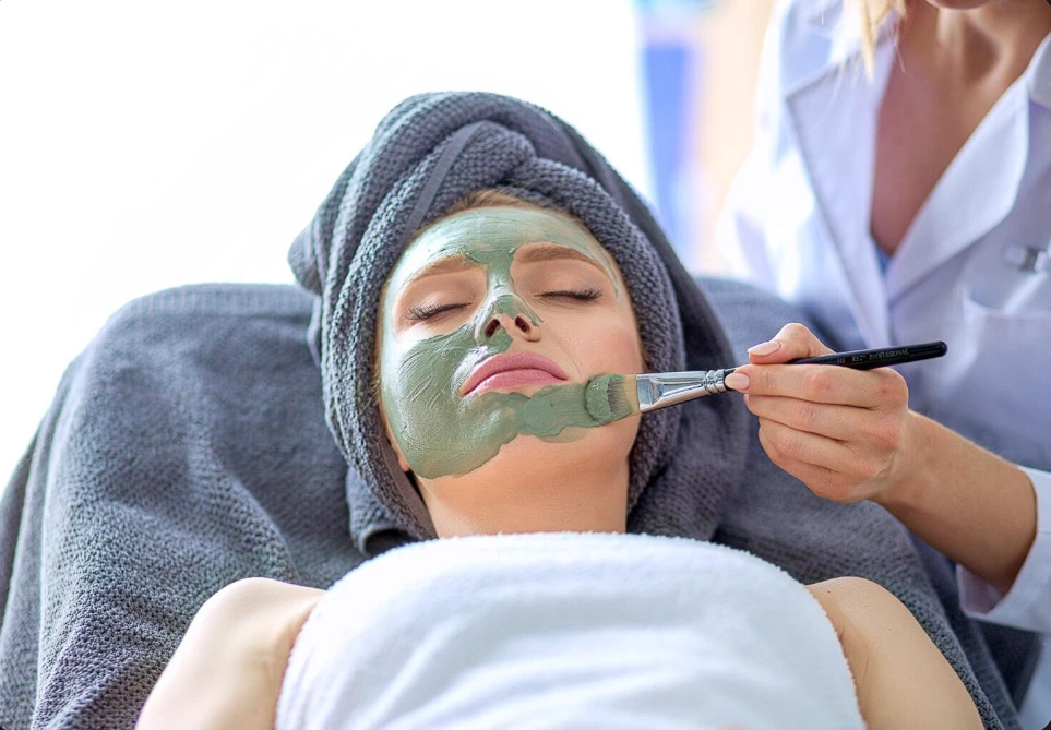 organic facial treatment in progress at ny brows and beauty salon, showing a client lying on a treatment bed with a white towel wrapped around their head while an esthetician applies a green, plant-based mask to the client's face using a brush. The treatment room has a calming ambiance with soft lighting and natural decor elements.