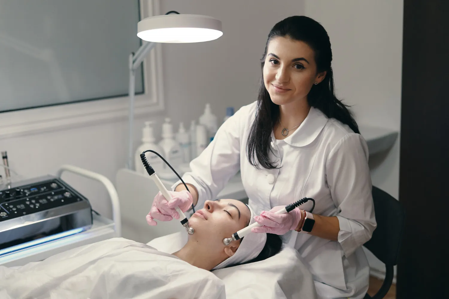 A smiling esthetician at ny brows and beauty salon in a white coat is performing a facial treatment on a client lying on a treatment bed. The esthetician is using handheld devices on the client's face, who has protective eye coverings and a hair wrap. Skincare products and equipment are visible in the background