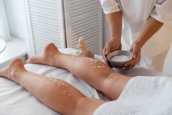 Close-up of a client's legs at ny brows and beauty salon receiving a body scrub treatment. A therapist's hands are applying a grainy scrub from a white bowl onto the client's legs, which are resting on a white towel. A wooden brush is visible on the bed next to the client's feet.